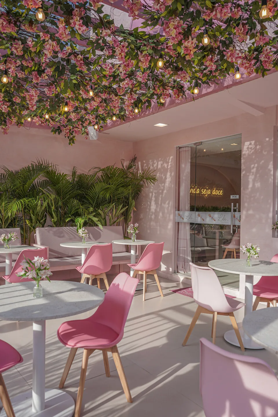 Varanda rosa sob copa de flores e lâmpadas, com mesas redondas, cadeiras rosa, palmeiras e a porta de vidro com neon ao fundo