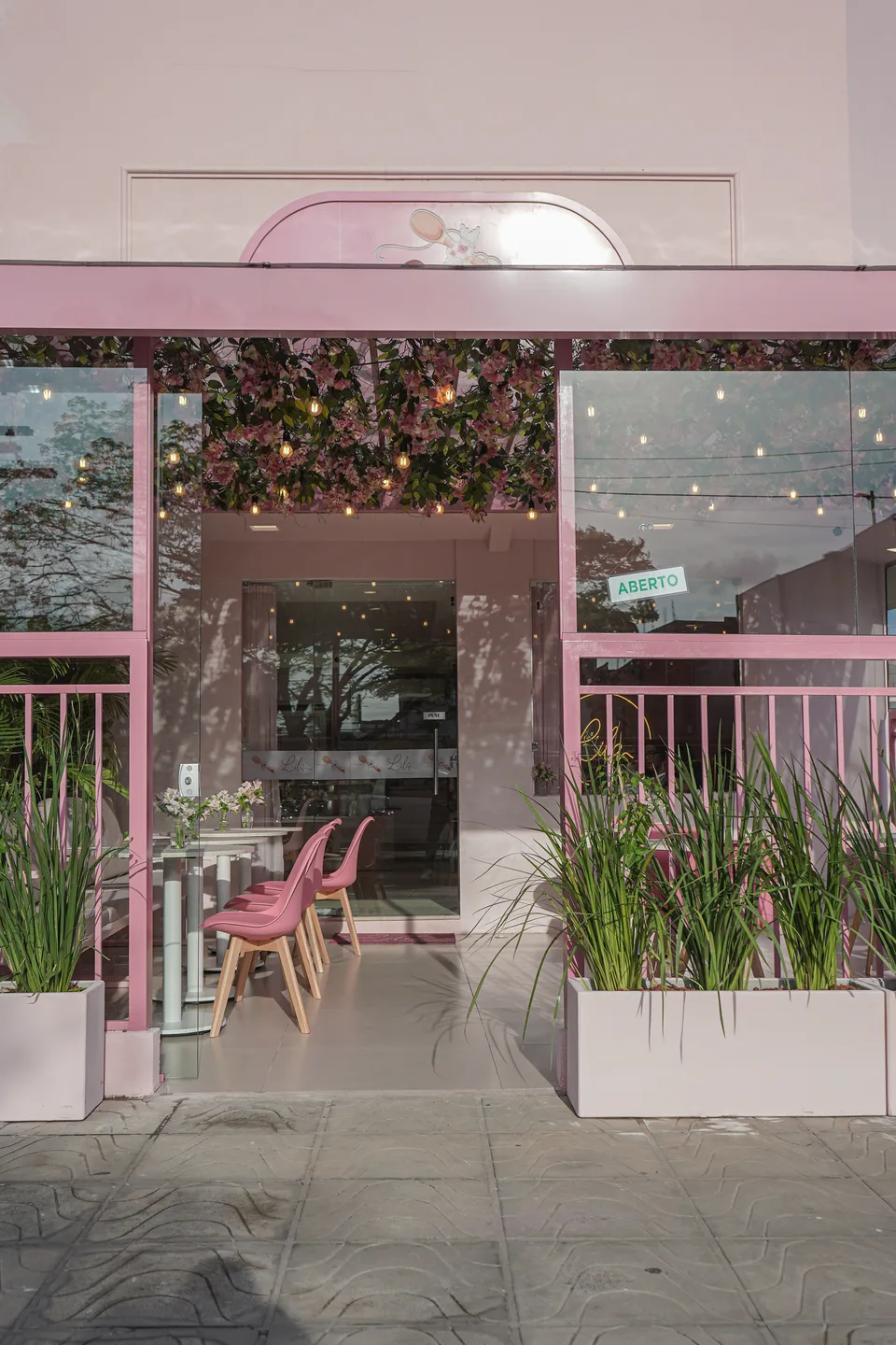 Fachada rosa com caixilhos de vidro, guarda-corpo de barras e floreiras brancas, revelando a varanda coberta de flores e a porta de entrada