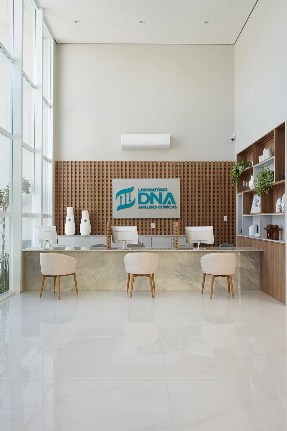Recepção de pé-direito duplo com fachada de vidro, balcão de pedra clara, cadeiras bouclé e painel em treliça