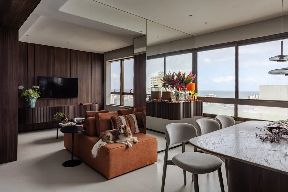 Estar integrado ao jantar, com sofá terracota, painéis de madeira escura, parede espelhada e janelas com vista para o mar