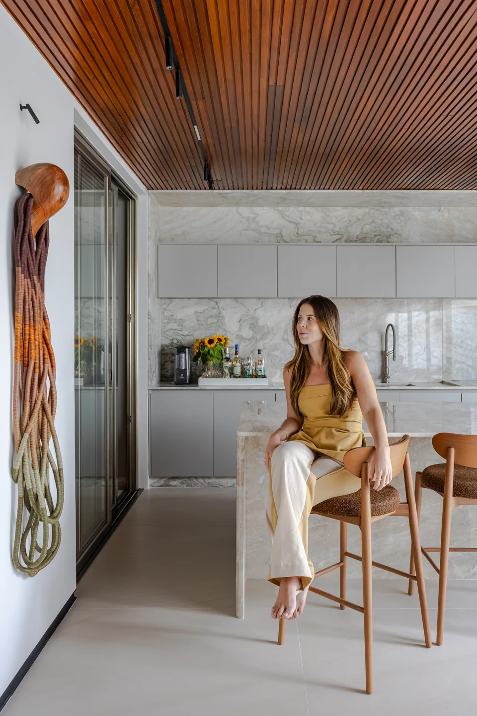 Carol Gomes sentada numa banqueta de madeira junto à ilha de pedra clara, numa cozinha com forro de madeira ripada