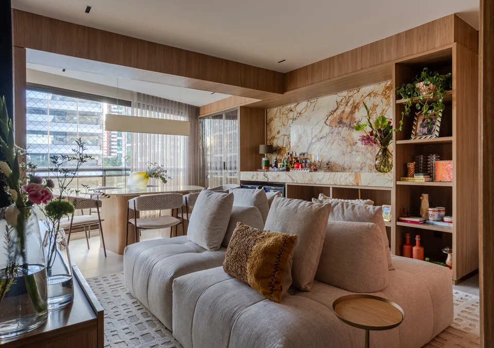Estar com sofá modular creme diante do bar em pedra veiada e estante de madeira, jantar junto à varanda envidraçada