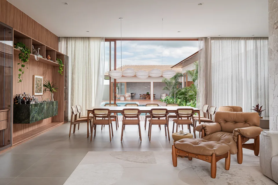 Estar e jantar abertos para o pátio da piscina, com poltrona de couro, mesa longa, bar em pedra verde e cortinas leves