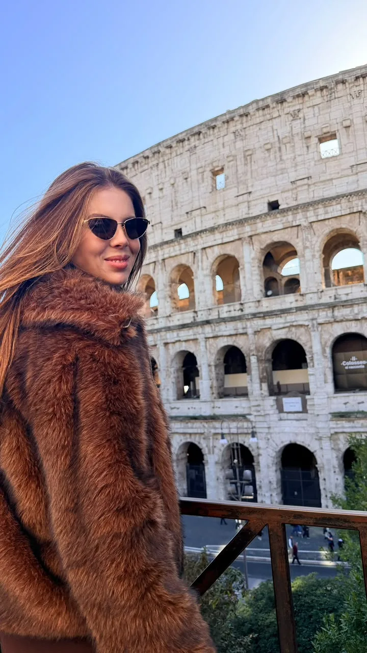 Carol Gomes de óculos escuros e casaco felpudo caramelo diante do Coliseu, em Roma