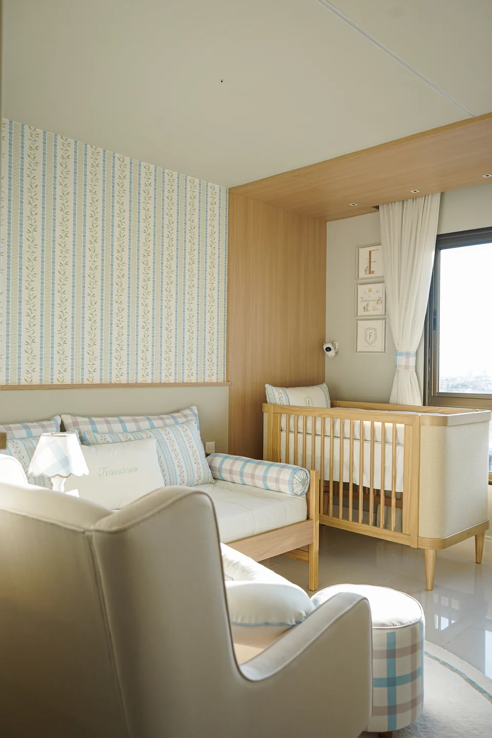 Quarto banhado de sol com cama sob papel listrado, berço em nicho de madeira junto à janela e poltrona creme