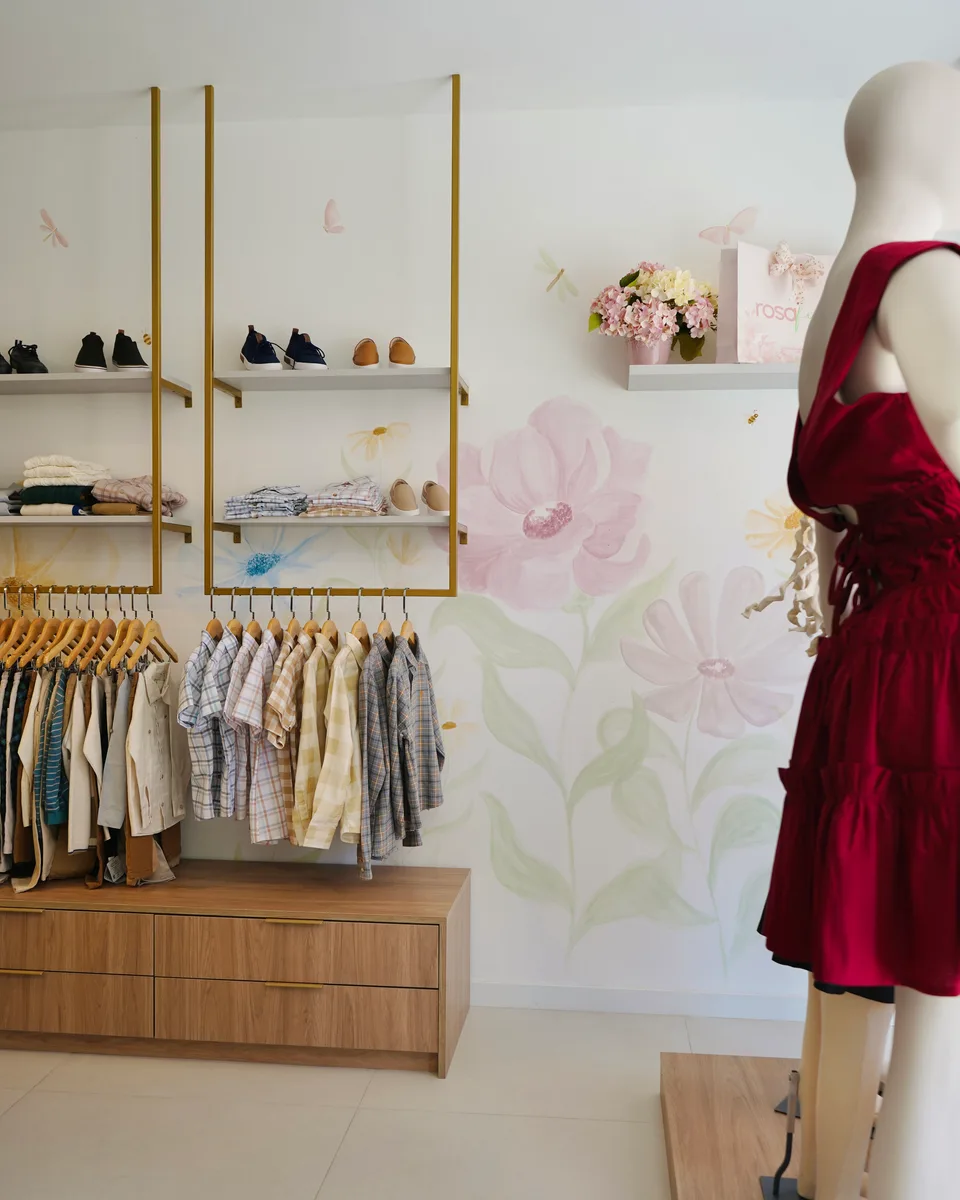 Molduras douradas suspensas com prateleiras de calçados e camisas, diante de grandes flores pintadas, e manequim com vestido vermelho