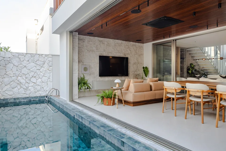 Piscina de pastilhas verde-azuladas junto à área gourmet aberta, com forro de madeira e muro de pedra